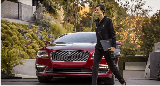 Woman walking past a red luxury car in a modern driveway, holding a tablet, surrounded by greenery.