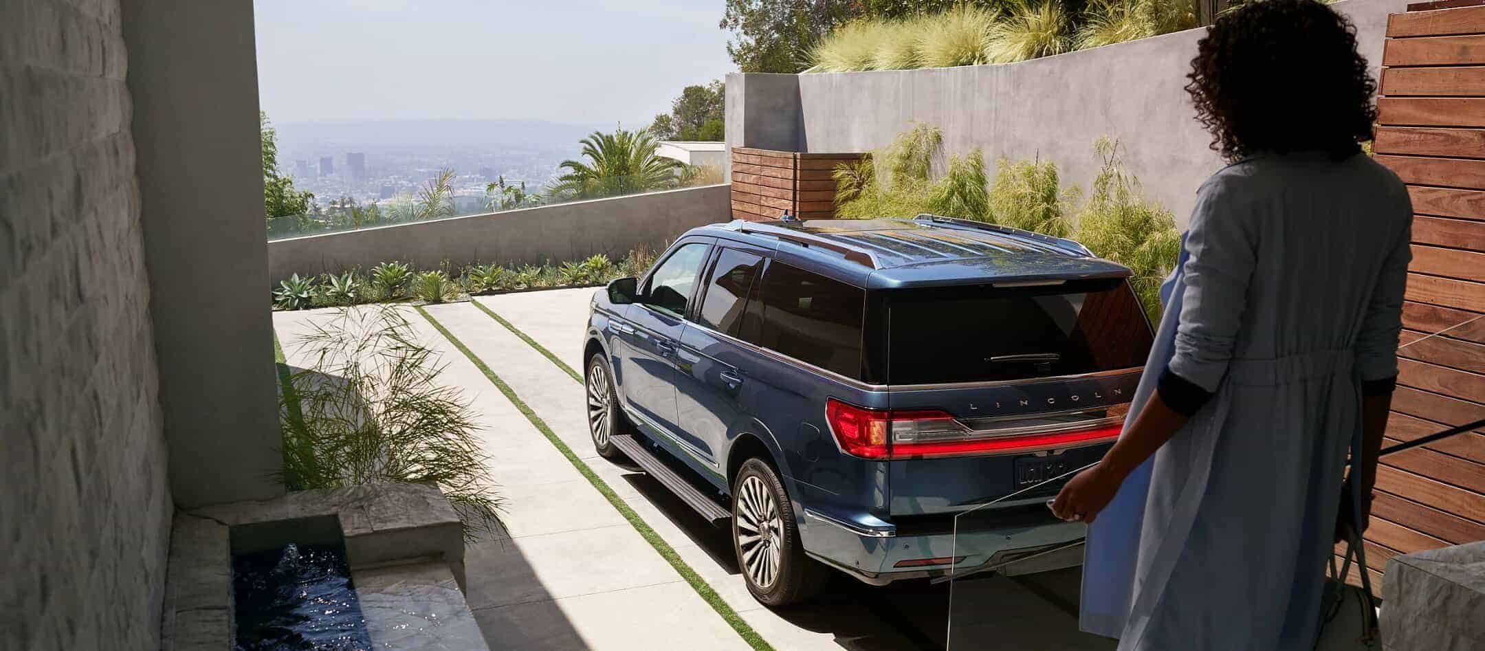 A person approaches a blue Lincoln SUV parked on a driveway with city views, surrounded by greenery.