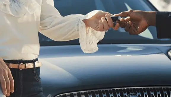 Two people exchange car keys in front of a vehicle, symbolizing a car transaction.