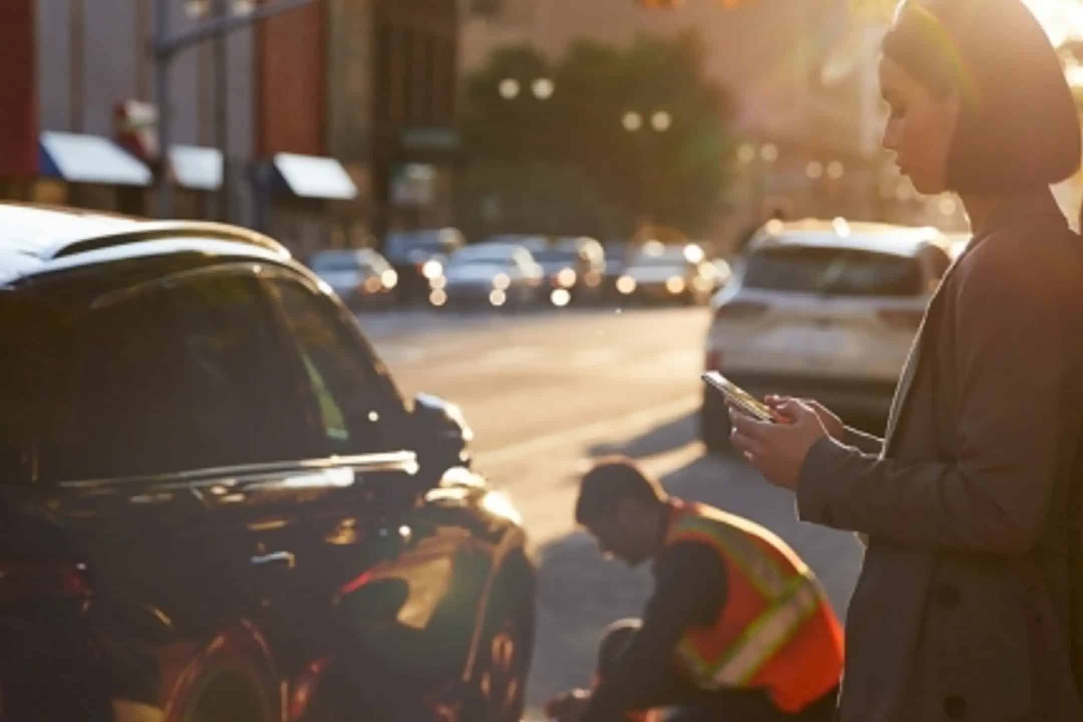 A woman uses a smartphone beside a street. Nearby, a worker in a reflective vest kneels by a car.