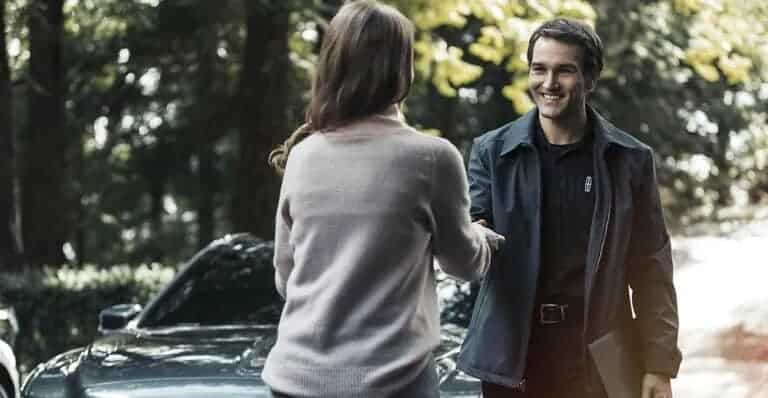 A woman and man smiling and shaking hands outdoors near a car.