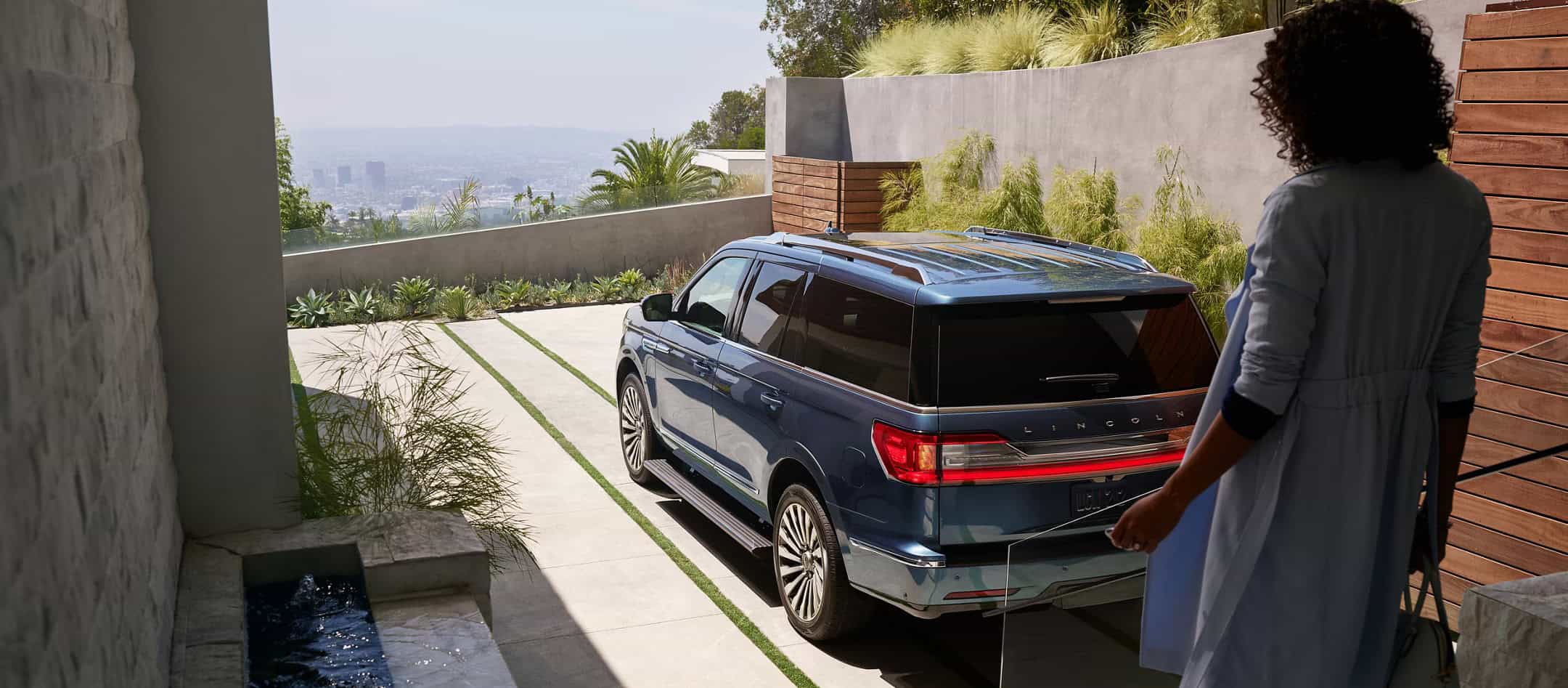 Woman approaching a parked blue SUV on a modern driveway with cityscape view.