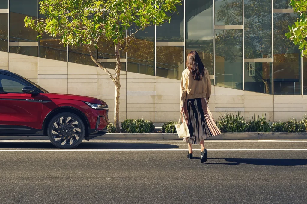 A woman walks on a crosswalk beside a red vehicle near a modern building with trees.