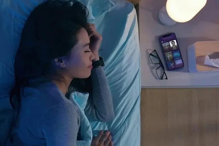 Woman sleeping with smart device on wrist; bedside table with phone, glasses, lamp, and tissues.
