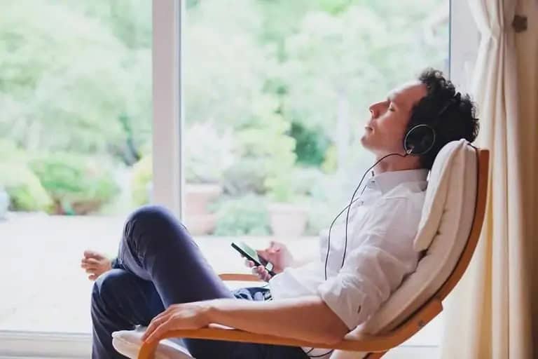 Man relaxing in chair with headphones, eyes closed, holding a smartphone, next to large window with garden view.