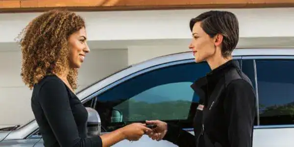 Two women exchanging car keys outdoors, standing beside a silver vehicle.