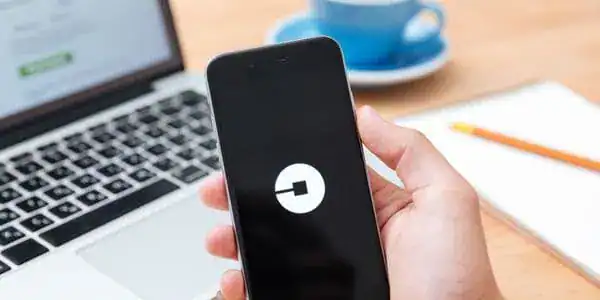 Hand holding smartphone with a ride-sharing app logo, next to a laptop and coffee cup on a desk.